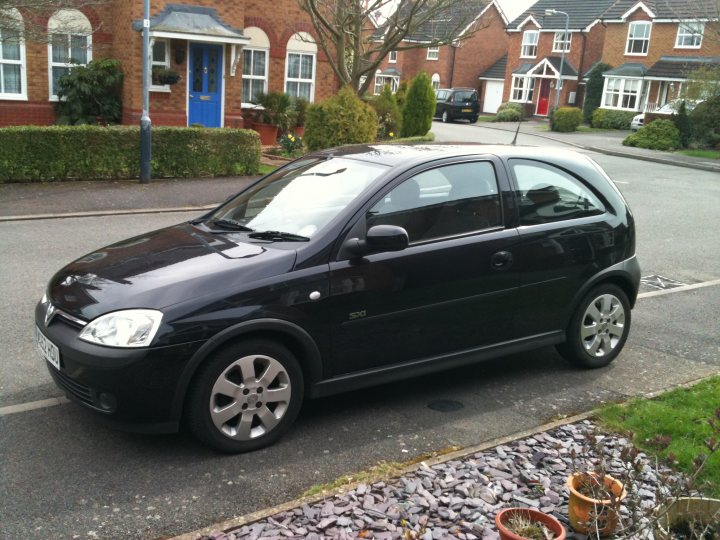 Pistonheads - The image shows a parked black compact car on a residential street. The car has a distinctive red headlight frame and a silver grille. Behind the car, there is a two-story residential house with a blue front door and a well-maintained garden featuring hedges and potted plants. The street appears to be quiet and the weather seems overcast, with a grey sky in the background.