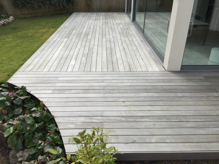 Decking sub frame - Page 1 - Homes, Gardens and DIY - PistonHeads - The image shows a section of a deck with vertical slats. The deck appears to be well-maintained with no visible signs of wear. It leads to a glass door, which reflects part of the indoor space. In the foreground, a plant with green leaves and purple flowers adds a touch of color and life to the scene. The background is slightly out of focus, but it seems to be a residential area with grass visible next to a fence.