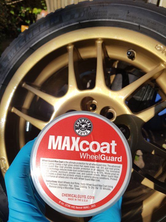 Pistonheads - The image shows a close-up of a wheel and tire assembly, likely for a vehicle. On the tire, there is a can of Maxcoat Wheel Guard, which appears to be a protective coating for tires. The can has a white label with red and black text, although the specific details on the label are not fully legible in the image. A small amount of the coating product seems to have been applied to the tire, visible as a red substance. In the background, there is another wheel with the same coating product applied to it. The setting appears to be an outdoor environment, possibly a driveway or a similar location where vehicle maintenance might occur.