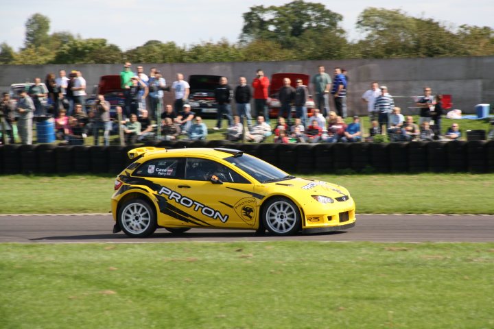 Saturday Castle Coombe Rally Day September Pistonheads - The image captures an exciting moment on a race track, where a yellow Proton sports car, adorned with a cheetah logo, is in motion. The cheetah logo on the side of the car suggests a brand association, likely indicating that it's participating in a branded event. The track is surrounded by several spectators, all attentively watching the thrilling action. The image conveys the adrenaline rush and competitive spirit typically associated with motorsport events.