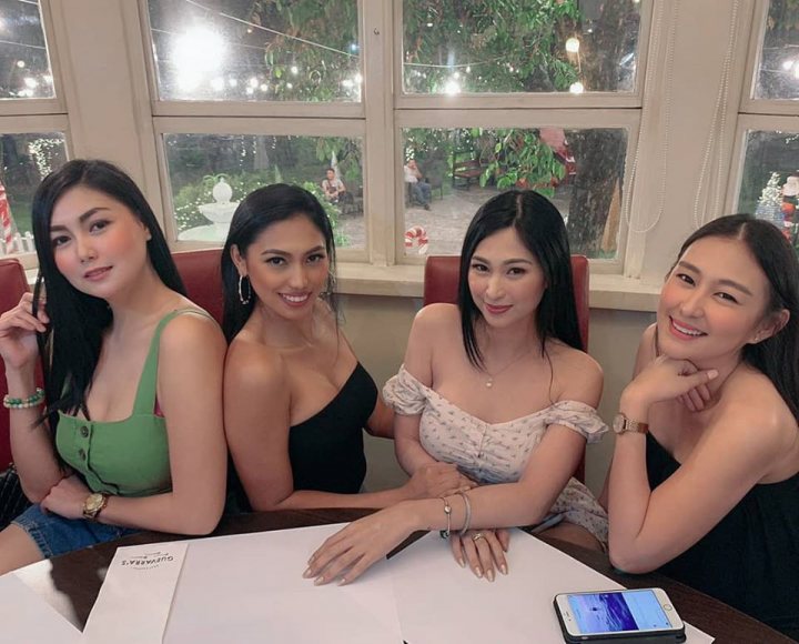 A group of people sitting around a table - The image captures a lively scene of four women posing for a photograph. They are seated around a dining table, which is adorned with various items including a cell phone and a cake. The woman on the left is holding a smartphone, while the other three women are smiling towards the camera. The backdrop reveals a restaurant setting, suggesting they might be celebrating or enjoying a meal together.