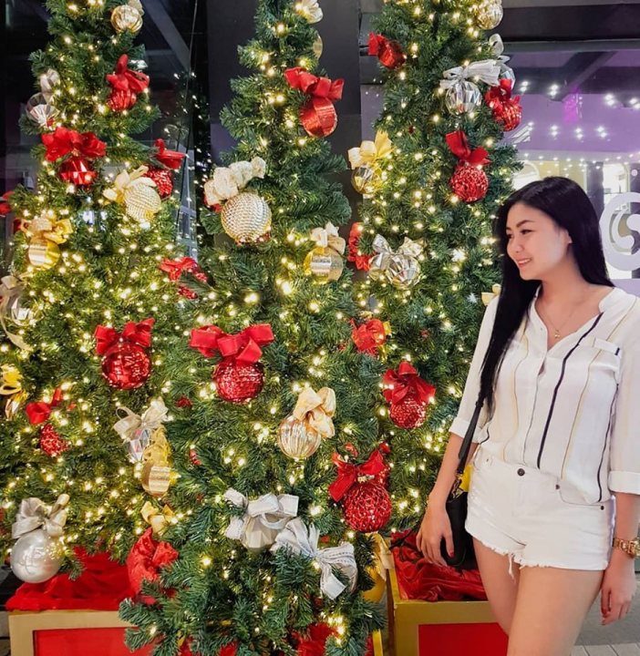 A woman standing in front of a table with flowers - In the image, a young woman is standing in front of a festively decorated Christmas tree. The tree is adorned with various decorations, including lights and red and gold ornaments. The woman is dressed in a white top and has a black handbag. She's posing for the photo, looking towards the camera with a smile on her face. In the background, there's another person, partially visible and also smiling at the camera.