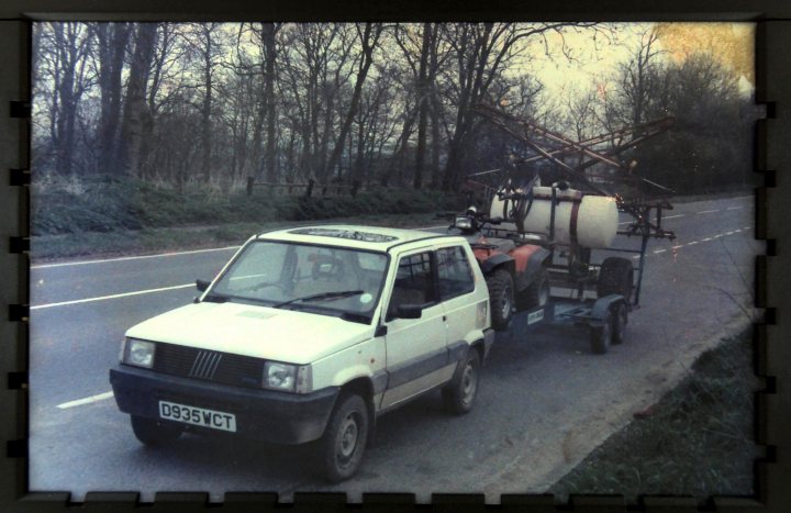 Fiat Panda 4x4 Mk 1 Fire.  A  Classic Car - Page 1 - Classic Cars and Yesterday's Heroes - PistonHeads - In the image, there is a vintage single-cab pickup truck with a white exterior pulling a trailer carrying a large, white structure that resembles a construction portal. The truck is driving on a city road, with trees and a fence visible in the background. The photo has a sepia tone, giving it a nostalgic feel, and the image's format appears to be a faded hardcover or a sketch. The truck is positioned in the center of the frame, drawing the viewer's attention to it. The trailer attached to the truck is connected securely, indicative of a purposeful and potentially job-related transportation.