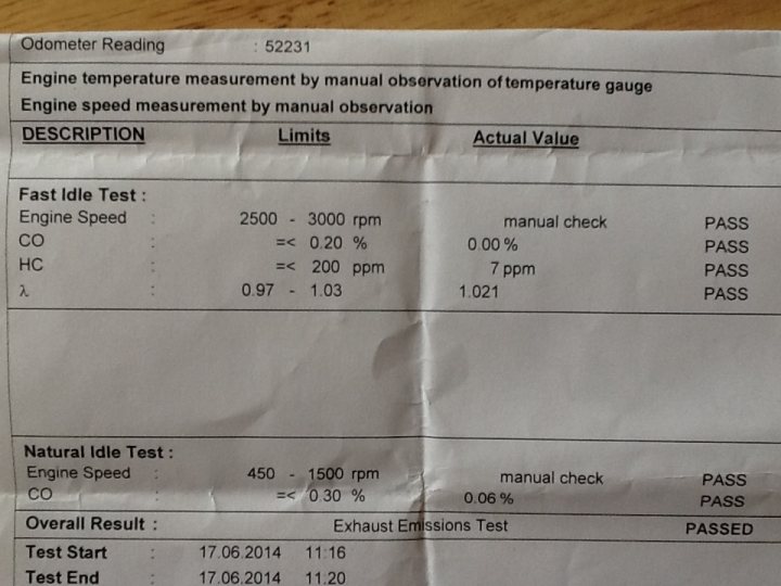 A close up of a sign on a wall - Pistonheads - The image is a photograph of a small sheet of paper or card with printed text and numbers that resemble data measurements. The document appears to be a tear-off section of a larger book that includes engine temperature measurement by manual observation and speed measurement by manual measurement, along with an actual value as a reference. Specifically, it details the engine's idling speed, measured with a CO gauge at the engine temperature. The test results show a reference natural idle speed of 450 rotations per minute (rpm) with the engine running at outdoor temperature, suggesting a calendar date of June 8, 2014, corresponding to the 863rd revision. The bottom right of the sheet includes a renewed ISO 9002 certificate number for an exhaust emissions test pass, indicating the time as 7:30 AM and the renewed certificate date as August 1, 2014. The text is somewhat blurred and the environment is indoors, with a wooden tabletop visible.