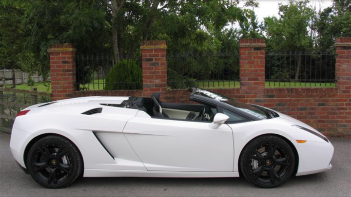 Newbie  - Page 1 - Gallardo/Huracan - PistonHeads - The image displays a luxurious white sports car parked on an asphalt driveway. The car features a distinctive and sleek design, with black rims and tires that contrast with the white body. It is a convertible model with the roof down, revealing two red brake lights at the back. In the background, there are trees and a brick wall that suggest a suburban setting. The photo seems to be taken during the day under clear skies, and the car is the focal point, occupying most of the frame.