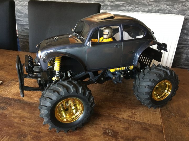 Show us your RC - Page 4 - Scale Models - PistonHeads - This image shows a toy car that is styled to look like a monster truck. The toy car is predominantly black with white accents and has oversized wheels and a raised suspension, which are characteristic features of real monster trucks. It's positioned on a table, surrounded by two other objects: a person in the background who appears to be wearing a hat and a pair of yellow gloves, suggesting they might have been handling the toy car or related items; and a smaller black car with large gold wheels, which could also be part of this collection. The scene suggests a casual indoor setting where someone has displayed their model cars or toys.