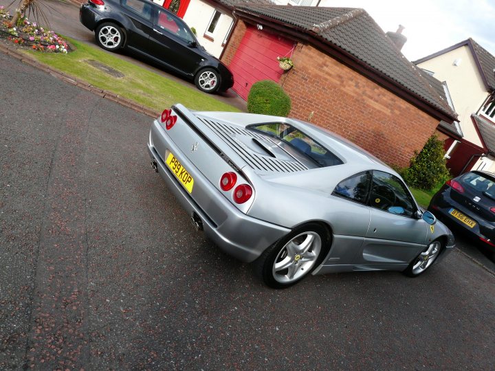 Pistonheads - This image features a gray sports car parked on a street with a brick house visible in the background. The car has red taillights and a yellow license plate, indicating that the vehicle is on the right side of the road. In the foreground, there is a flower plant adding a touch of greenery to the scene. There are also other cars parked along the street, suggesting a residential area. The sky is visible in the top right corner of the image, indicating that it might be late afternoon or evening, given the warm light and shadows cast.