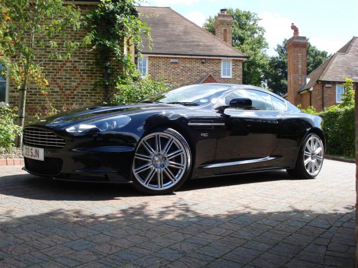 Like new again - Page 1 - Aston Martin - PistonHeads - The image showcases a sleek black Aston Martin sports car parked on a brick driveway. The car is the main subject, with its shiny black exterior gleaming under the sunlight. It is positioned somewhat sideways, giving a clear view of its wide stance and chunky wheel arches. The brick driveway leads the eye towards the house in the background, which sports a classic red brick facade. The sky above is blue and clear, suggesting a pleasant day.