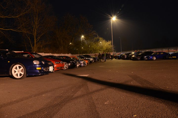 Pistonheads March Essex - The image depicts a parking lot at night, filled with various cars and a motorcycle. There is a person standing near the front of the photo. A few lights from the vehicles illuminate the scene, and the darkness around suggests it might be late at night. A fence can be seen in the upper right corner of the photo, possibly indicating the boundary of the parking lot.