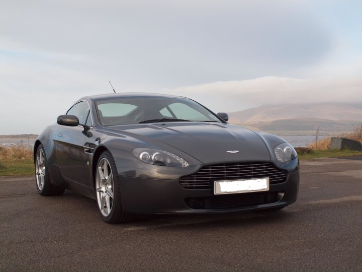 Another New owner - Page 1 - Aston Martin - PistonHeads - The image features a sleek black Aston Martin sports car elegantly parked on a smooth, asphalt road. The car's glossy surface reflects the light, highlighting its muscular silhouette. In the backdrop, a vast, open landscape stretches out, dotted here and there with trees and mountains under a clear blue sky. The precise positioning of the car on the road, coupled with the flared fenders, enhances the car's aggressive stance. The overall scene suggests a serene drive through scenic countryside, with the Aston Martin as the centerpiece of this picturesque setting.