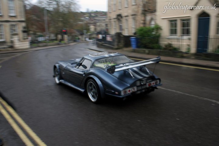 Sunday April 25th Bradford on Avon - Marocs - Page 1 - South West - PistonHeads - The image depicts a dark blue race car with a distinctive aerodynamic design, including a large rear spoiler and side-mounted aero scoops. The car is traveling down a street with a characteristic road surface that creates double yellow lines, indicating a no-passing zone. The setting appears to be urban, with buildings and a traffic light visible in the background, suggesting the photo may have been taken in a European city. There is a watermark text overlaid on the image, which might indicate the name or logo of the owner or photographer.