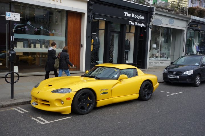 Black OEM wheels - Page 1 - Vipers - PistonHeads - The image captures a vibrant city scene on a busy street. The street is lined with storefronts and shops, each one distinct and inviting. In the foreground, the street is dominated by two yellow cars parked neatly along the curb. One of the yellow cars is a convertible car sports car, adding a touch of luxury to the urban setting. Pedestrians can be seen casually strolling along the sidewalk, adding life and movement to the scene. A parking meter stands tall next to the parkable car, marking the boundary for parking in this area. The black awnings over the storefronts provide a stark contrast to the blue sky above, creating a dynamic atmosphere characteristic of city life.