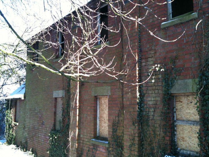 Nsomerset Ruins Pistonheads - The image shows a brick building with numerous windows, some of which appear to be boarded up. A tree with bare branches is in the foreground, suggesting it might be autumn or winter. The perspective of the shot is such that part of the image is slightly off-center, adding a sense of depth to the photo. The old and abandoned appearance of the building, with its bare tree and overgrown vines, conveys a sense of solitude. The overall mood of the image is somber and gloomy, with the monotony of the brick walls punctuated by the stark contrast of the bare surroundings.