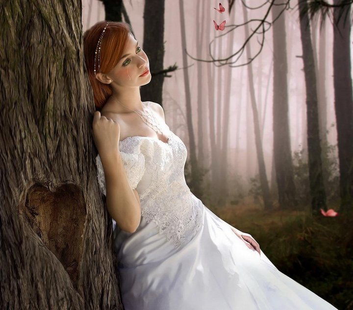 Girl Art Dress - In this enchanting image, a woman clad in a white wedding dress is seen posing against a tree adorned with a heart-shaped decoration. The setting is a serene forest with delicate pink blossoms gracing the ground. The scene is complemented by a romantic atmosphere, further enhanced by ethereal butterflies flying around her silhouette. The bride's location against the gnarled tree and the scattering of blossoms on the ground create a picturesque moment of celebration and connection to nature.