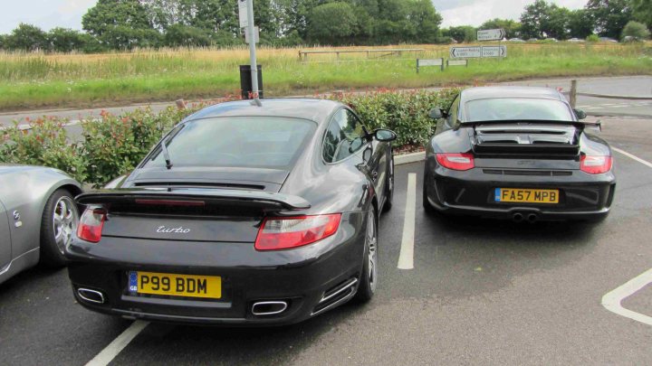 Hare Breakfast Meet - August - Page 1 - Kent & Essex - PistonHeads - The image depicts a tranquil scene in a parking lot. Two distinctive cars, a black sports car and a silver Bentley Flying Spur, are parked side by side. Both cars are marked with yellow license plates and are positioned on a spot with white lines, indicating parking regulations. Additionally, the sports car proudly displays the name "Torroti" on its back, perhaps indicating the dealership or car customization service. The background is slightly blurred but suggests a peaceful park setting, adding to the overall calmness of the scene.