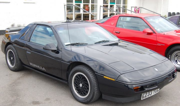 What American car for a UK garage? - Page 1 - Yank Motors - PistonHeads - The image shows two cars parked side by side. The car on the right is predominantly red, featuring a convertible design with an open-top. On the left is a car with a black body and a black soft-top roof, modeled after a Porsche Cayman. Both vehicles have visible headlights and black wheels. They are parked on a surface that seems to be a parking lot, with an entrance in the background that has an awning.