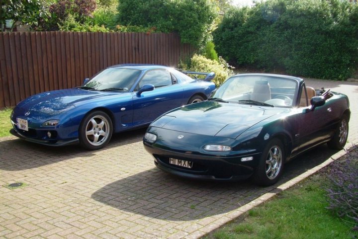 Best 2 Car Combos - Page 3 - General Gassing - PistonHeads - The image showcases an outdoor scene where there are two cars parked side by side on a brick-paved driveway. The car on the left is a dark blue sports car with the top down. The car on the right is a sleek, black sports car with the top up. Both cars appear to be well-maintained and showcase the characteristics of sports cars, with streamlined bodies and sporty profiles. They are parked against a backdrop of verdant greenery, creating a contrast between the man-made objects and the natural environment.