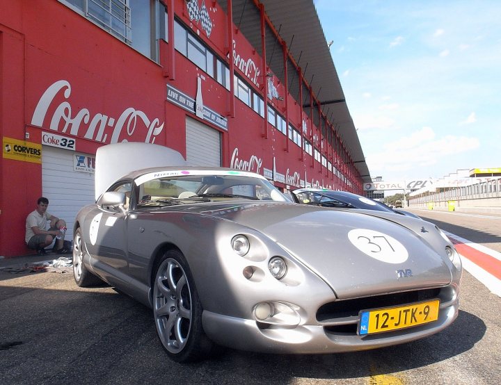 Pistonheads Oil Filler Cap - The image presents a striking scene on a red brick street. Dominating the frame is a silver, vintage Jaguar, parked and facing us. A man can be seen leaning casually against the Jaguar's hood, adding a touch of human element to the scene. In the background, a vibrant red and white Coca-Cola advertisement grabs attention, its white lettering standing out against the bold backdrop. Further back, another car can be spotted, parked and facing away from the camera. The scene is framed by a building with a red awning and a green corrugated roof, providing a colorful contrast to the red brick street.