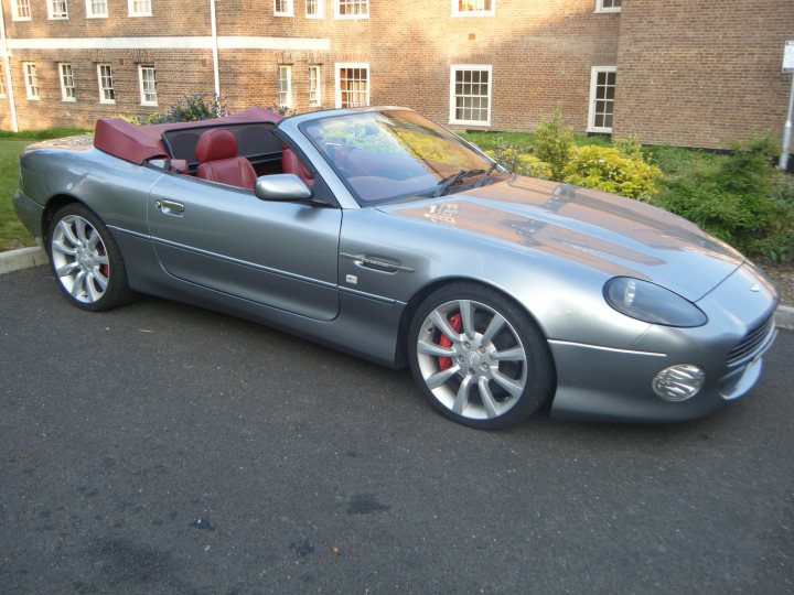 Your DB7 pictures. - Page 1 - Aston Martin - PistonHeads - The image features a sleek, two-door convertible car parked on a driveway. The car has a shiny silver exterior, a red interior, and its top is down. The vehicle is positioned at an angle that shows off its side view and the curvature of the road. The setting appears to be a suburban area with a brick house, garden, and tree visible in the background.