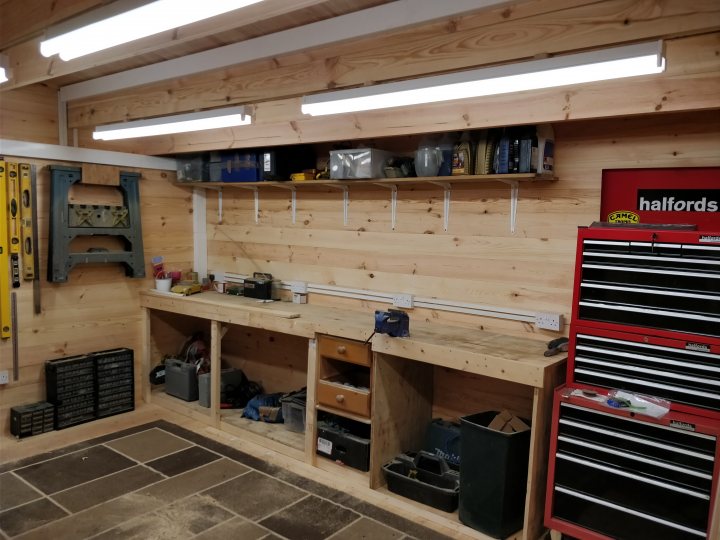 garage lighting ideas - Page 1 - Homes, Gardens and DIY - PistonHeads - The image depicts a neat and organized garage or storage room. There is a prominent bench with shelves mounted above it. The wooden walls feature a half-boards design at the top. Multiple precise tools and accessories, including cords, visor, wrenches, and others, are well-organized on the shelves. On the right side, a red tool chest stands against the wall. The environment suggests a space dedicated to work or repairs.