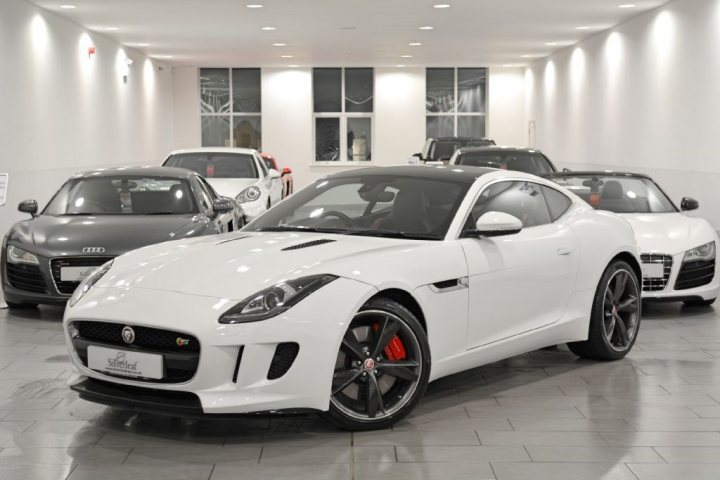 I've just bought an F-Type (pictures) - Page 1 - Jaguar - PistonHeads - The image showcases a dealership with modern and elegant interiors. The focus is on one white sports car in particular, highlighted by its glossy finish and featuring characteristics typical of high-performance vehicles. The car's design is sleek and streamlined, suggestive of top-tier luxury brands. Around the car, two other models of a smaller size are displayed, with their positioning offering a comparison of size and design. Outside the showroom, the area is currently empty, enhancing the attention drawn to the cars.