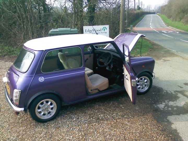 Pistonheads Mayfair Mini - The image shows a small, purple car parked on a gravel surface. The car's hood is lifted up, revealing the engine beneath. The car features a distinctive white stripe on its side. In the background, there's a sign for a cafe, indicating that the car is in a rural or less populated area, possibly the countryside. The overall scene suggests a day trip or a stop during a longer journey.