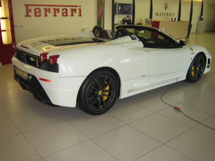 Pistonheads Interest - The image features a white Ferrari sports car parked in what appears to be a showroom or auto dealership. The car is oriented towards the right side of the frame, showcasing its front and side profile. The vehicle's design elements such as the rear spoiler and impeller grille are prominent. It is equipped with dual black rims and yellow brake calipers. On the wall behind the car, the 'Ferrari' logo is displayed. The quality of the image suggests it might be used for promotional purposes or commercial inventory.