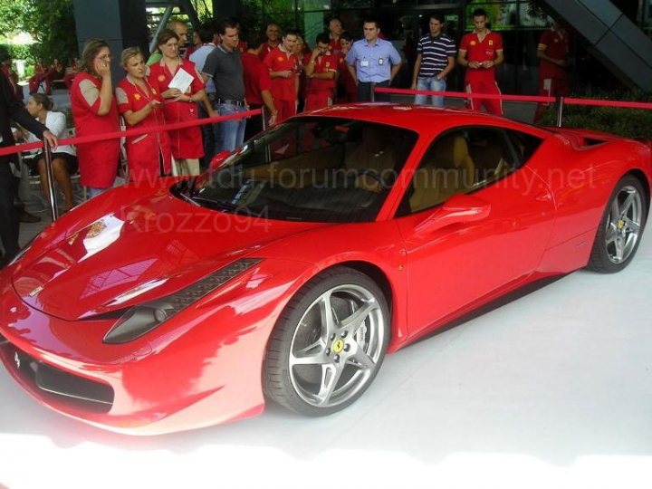 Italia Ferrari Pistonheads Ive - The image showcases a vibrant red Ferrari sports car parked on a white surface. A crowd has gathered around the exclusive car, with people in various outfits standing behind a red rope barrier. The individuals appear to be observing the Ferrari, possibly admiring its sleek design and polished finish. The scene suggests this could be a promotional event where the Ferrari is the center of attention. The environment seems to be an open area with a black steel structure in the background.