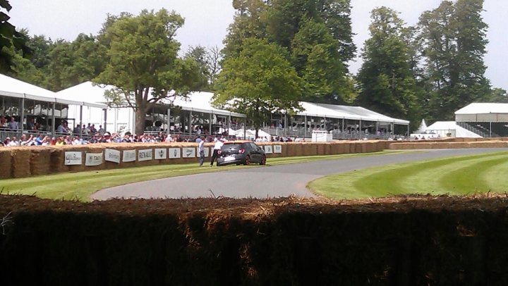 Goodwood MMS / FoS 2012 Pictures - Page 1 - Goodwood Events - PistonHeads - The image captures a vibrant scene of an outdoor event, possibly a race given the presence of a track. A crowd of spectators are gathered around the track, engaged in various activities and enthusiasm. There are numerous umbrellas, likely standing as stalls or providing shade from the sun. Surrounding the track is a fence constructed from various materials, and in the distance, several tents and stalls can be spotted, contributing to the bustling atmosphere. There is a single black car driving on the track, possibly a pace car or a participant's vehicle.