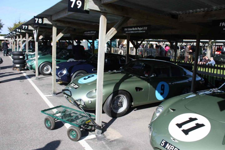 Revival images - Friday  - Page 1 - Goodwood Events - PistonHeads - In the image, a line of vintage cars are parked parallel to each other under wooden beams supported by metal poles. The cars are neatly arranged, their antique designs and polished exteriors gleaming under the sunlight. The wooden canopy provides a vintage aesthetic to the scene. In the background, people can be seen walking around, possibly admiring the collection of classic cars. The atmosphere seems like a car show, with the cars being the main attraction. The overall scene suggests a day of leisure filled with car enthusiasts appreciating the beauty of vintage automobiles.