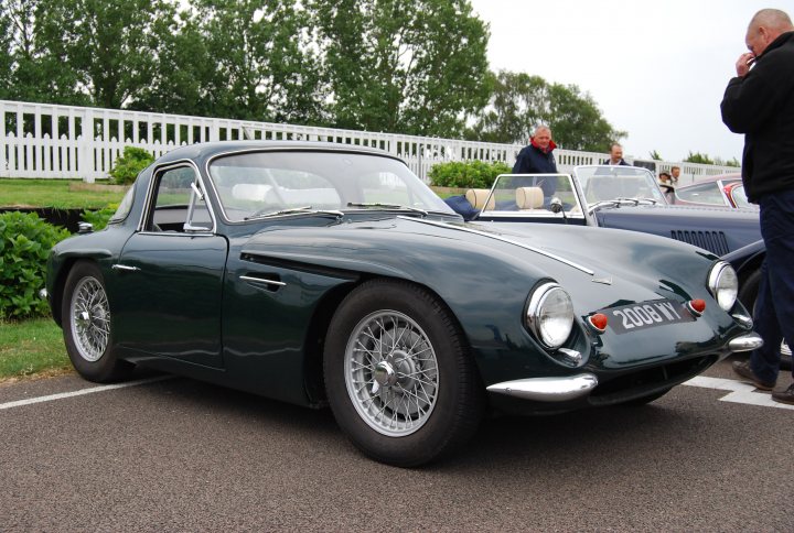 Early TVR Pictures - Page 4 - Classics - PistonHeads - The image captures a vintage scene of threefold transportation. A green vintage car is the centerpiece, elegantly parked on a paved surface. In the background, a stylish man wearing sunglasses and a black jacket adds a touch of glamour. The presence of a blue car and additional people in the scene suggests a casual gathering or event.