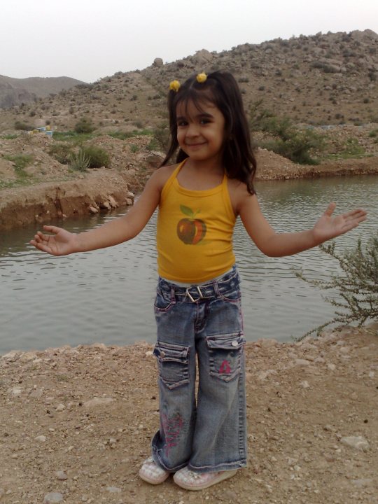 In the image, there is a young girl standing on a rocky surface with a body of water visible behind her. She has long dark hair and is wearing a yellow tank top with an embroidered apple. She's also sporting denim jeans rolled up to her calves. The girl is touching her arms with her hands and has a wide smile on her face, giving the impression of joy or surprise. The overall setting appears to be outdoors, possibly in a desert-like environment due to the arid land and absence of dense vegetation.