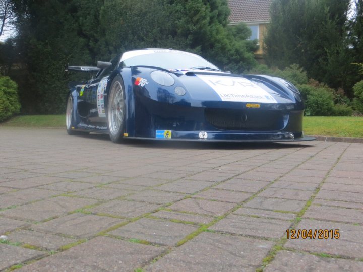 Built in Bruntingthorpe  - Page 1 - Noble - PistonHeads - The image shows a sports car parked on a brick-paved surface surrounded by greenery. The car, with its aerodynamic design, features a large front grille and a prominent racing-style spoiler on the rear. It appears to be a custom or rally-style car, and there's a sticker around the windshield, possibly indicating a racing team or event participation. The environment suggests a quiet suburban neighborhood.