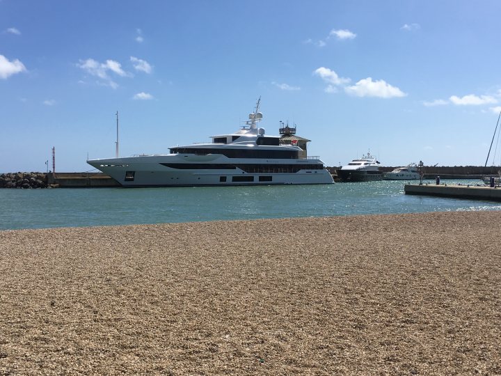 super yachts 60million+ - Page 167 - Boats, Planes & Trains - PistonHeads - The image is a color photograph showcasing a large, luxurious yacht with a white hull fully docked at a marina. The yacht features multiple levels, numerous windows, and external deck space. It seems to be a clear day with a calm sea visible in the background, indicating favorable weather conditions. There are no visible people or activity within the marina that can be confidently identified from this perspective.