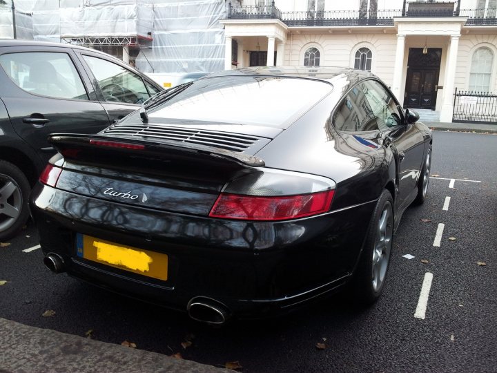 GT1 block owners - catastrophic coolant leak prevention - Page 1 - Porsche General - PistonHeads - This image shows a rear view of a black turbo Porsche parked on the side of a street. The car is sleek and black, with a prominent tail pipe and a yellow license plate that reads "TRU". The pavement appears to be wet, and a building with an imposing black gate looms in the background. Two other cars are visible in the background, one parked behind the Porsche and the other partially visible on the opposite side of the street. The street has white markings, indicating a designated parking lane and traffic lanes.