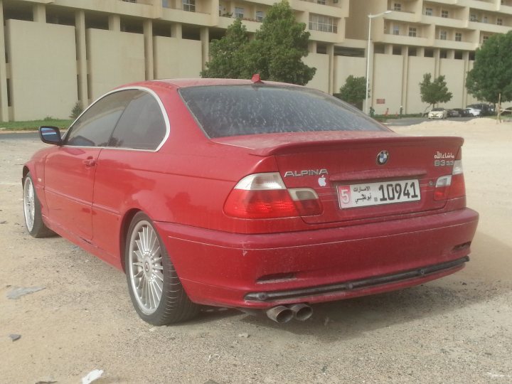 CARS IN DUBAI WHICH NEED SAVING!   - Page 1 - Middle East - PistonHeads - The image captures a small red sports car parked on a beige sandy area. The car appears to be a BMW, identifiable by the badge on the trunk and the distinctive styling. The car's bodywork is covered with a dark substance, possibly a protective layer or tar spots, which gives it a shiny look. The car is positioned against a backdrop of residential buildings, adding an urban touch to the otherwise desert-like setting. The car's wheels and the exhaust pipes are prominent features in the scene. The lighting suggests it might be a midday shot, enhancing the details on the car and in the background.