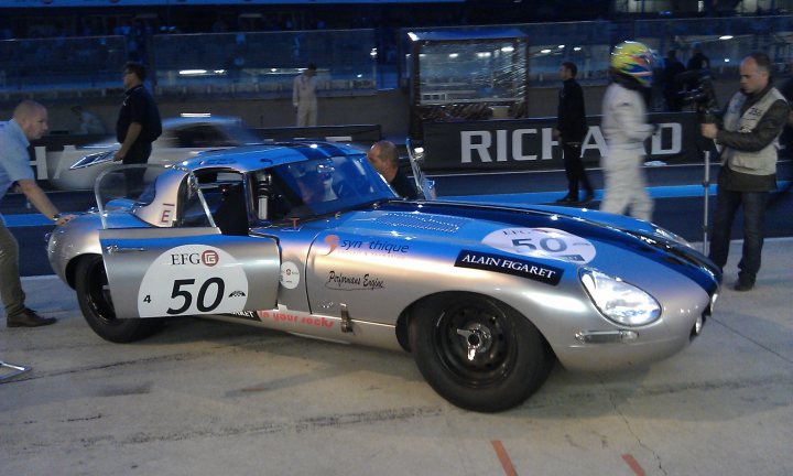 A car is parked on the side of the road - Pistonheads - The image is a photograph set in what appears to be a garage or pit area at a race track. It features a blue race car with a number "50" displayed prominently on its side, along with sponsor logos and the word "Alain". Near the car, there are a handful of individuals standing around, including two people walking past each other in the background. Most of the individuals are dressed in casual clothing, suggesting they are not participants in the race. The attire of one person near the car, however, seems more appropriate for a race context, hinting at their professional role in the event, possibly related to driving or logistics. The overall atmosphere of the image conveys the anticipation and activities typically associated with a racing event.