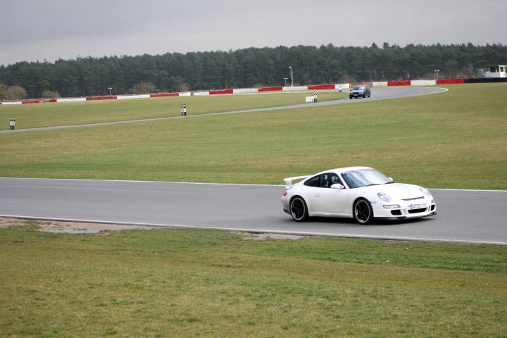 Track day at Snetterton Friday 15th February - Page 1 - East Anglia - PistonHeads - The image is a color photograph showing a white sports car moving on a race track with several lanes. The car is a two-door coupe with a sleek design and prominent rear wing, indicative of a sports or racing vehicle. It appears to be mid-turn, leaning into a curve on the course. The track is enclosed by a grassy area, and in the distance, other vehicles, including one that seems similar to the lead car, can be seen following the same path. The lighting suggests it might be an overcast day, with diffuse light casting soft shadows on the ground and vehicles.