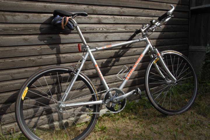 Any fixie fans on here ? - Page 2 - Pedal Powered - PistonHeads - This image showcases a single-speed bicycle parked against a wooden wall or fence. The bicycle is currently unridden and leans against a wall which may be made of bamboo or a similar material. It appears to be a casual, outdoor setting. The bicycle is equipped with orange and white pedals, adding a pop of color to the silver frame. There are no visible texts or logos indicating any specific models or brands. It's a clear day with no people or moving vehicles in the immediate vicinity, creating a peaceful and serene atmosphere.