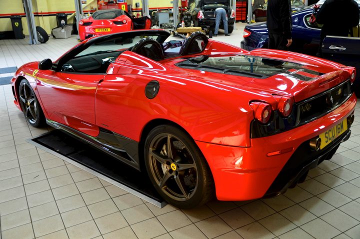 16M - Page 1 - Ferrari V8 - PistonHeads - The image depicts a red Ferrari car parked in a workshop or store area, where there are other vehicles. This vehicle is showcased from the front with its hood up, revealing the engine. There are several attendees present, seemingly observing or maintaining the cars. The cars, including the Ferrari, are positioned on elevated platforms, and the space appears to be well-lit and professionally maintained. The presence of the Ferrari and the attentive attendees suggests an event or professional setting focused on high-end vehicles.