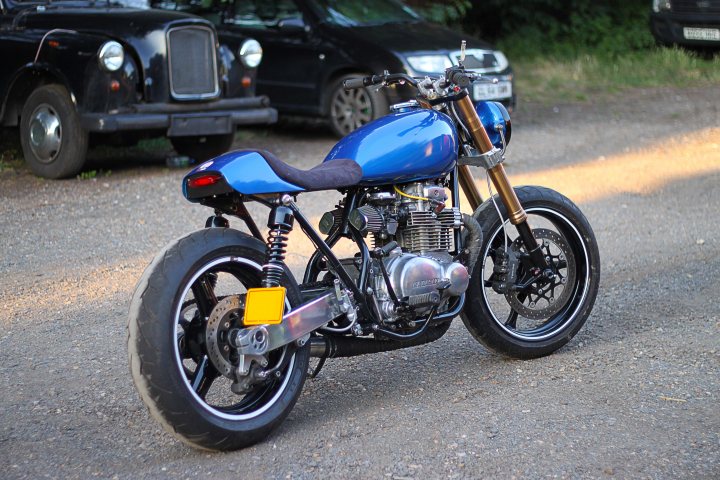 Show your 70's bike... - Page 5 - Biker Banter - PistonHeads - The image captures a scene on a gravel road, where a vibrant blue motorcycle stands as the main focus. The motorcycle, parked facing the camera, leans slightly to the left as if poised to ride off at any moment. It features a black seat, a large cylindrical engine, and a yellow license plate. In the background, two cars can be seen parked further down the road, adding depth to the image and suggesting a remote location. The overall setting evokes a sense of tranquility and freedom, perhaps hinting at the motorcyclist's journey ahead.