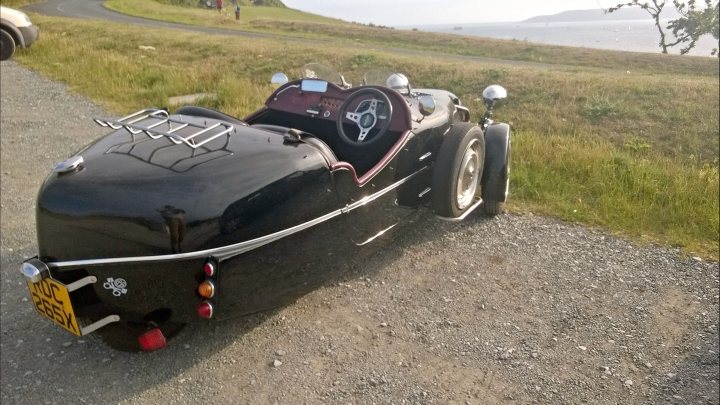 LOMAX 223 - slow max the right kinda trike - Page 5 - Readers' Cars - PistonHeads - The image shows a unique, two-wheeled vehicle parked on a gravel surface. This vehicle combines features of a motorcycle and a small car: a pair of rear-mounted wheels, a single front wheel, a small closed enclosure that could be a cockpit, and a windshield shielding the cabin. The bodywork is predominantly black with red accents, and there is a yellow license plate. In the background, there is a verdant landscape with a hint of a body of water, giving the impression of a leisure setting. The vehicle appears to be ready for travel, sitting motionless on the left side of the frame.