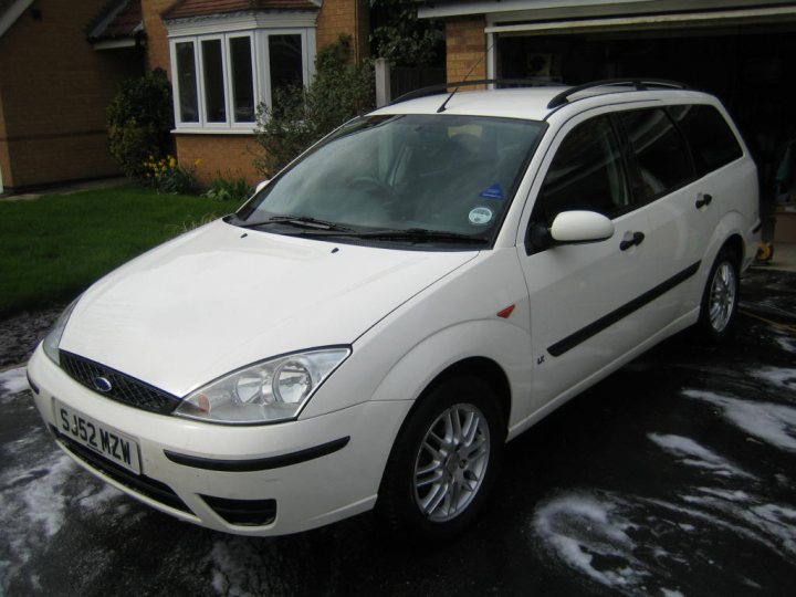 Pistonheads Megamile - The image features a close-up of a white Ford Mondeo parked in a driveway. The car is facing forwards, and its windows and side mirrors are clearly visible. Behind the Ford, a portion of a brick house and a window with black trim can be seen. The surface of the driveway appears to be wet, suggesting recent rainfall. The overall lighting and shadows indicate it might be a cloudy day, and the image is taken outdoors during the day.