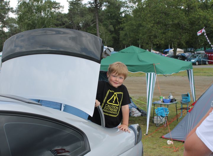 Le Mans  2012 ..... Pics and Storys - Page 5 - HSV & Monaro - PistonHeads - In the image, a young boy is standing in the front of an open car hood, smiling broadly as he peers out. The car, a sleek silver model, is parked in a camping area, as indicated by the surrounding tents and camping gear. The background reveals a busy camping scene with multiple vehicles and other outdoor enthusiasts. The boy's bright attire contrasts with the more subdued colors of the camping equipment. The overall atmosphere of the image is cheerful and exploratory.