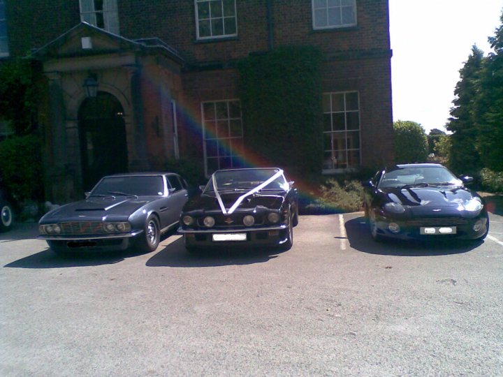 How about an Aston wedding car photo thread? - Page 1 - Aston Martin - PistonHeads - The image shows a row of three antique cars parked in an outdoor lot in front of a brick building with a variety of glass windows. The cars are sleek with aerodynamic designs, featuring compact and curved bodies, and they are parked closely together, occupying all of the visible parking spaces. The vehicles have a classic appearance, reminiscent of the 1960s or 1970s, with minimal embellishments and a focus on smooth lines and stylish design. The scene is bathed in natural light, indicating a clear day or sunny conditions.