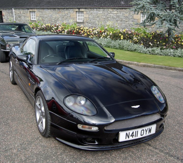 Astons & Ferraris at Glenfiddich distillery - Page 1 - Aston Martin - PistonHeads - The image captures a scene on a street where a sleek, new black convertible car is parked. The car's polished exterior and shiny rims gleam under the daylight. Its soft-top roof is down, revealing a tidy interior filled with various controls and features. The car's license plate appears to be from the United Kingdom. The serene setting is highlighted by the presence of a stone building and well-maintained hedges in the background.