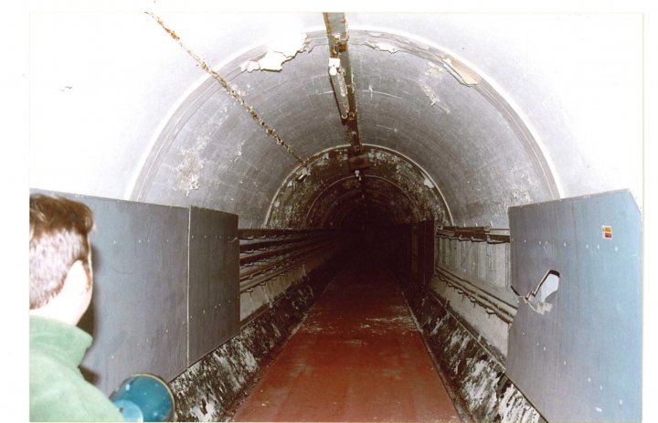 Housewith Pistonheads Difference - The image depicts a person standing in a dimly lit, underground corridor or tunnel. The person appears to be holding a blue object that resembles a block, connecting two walls or structures in front of them. The tunnel is curved and exhibits some unpainted or worn areas, suggesting age or multiple uses. The walls within the tunnel are not maintained and are made of concrete, showing exposure to water or moisture. There is no visible person on the platform, which could further imply the location is not currently in use, or the uploaded image does not show a busy area.