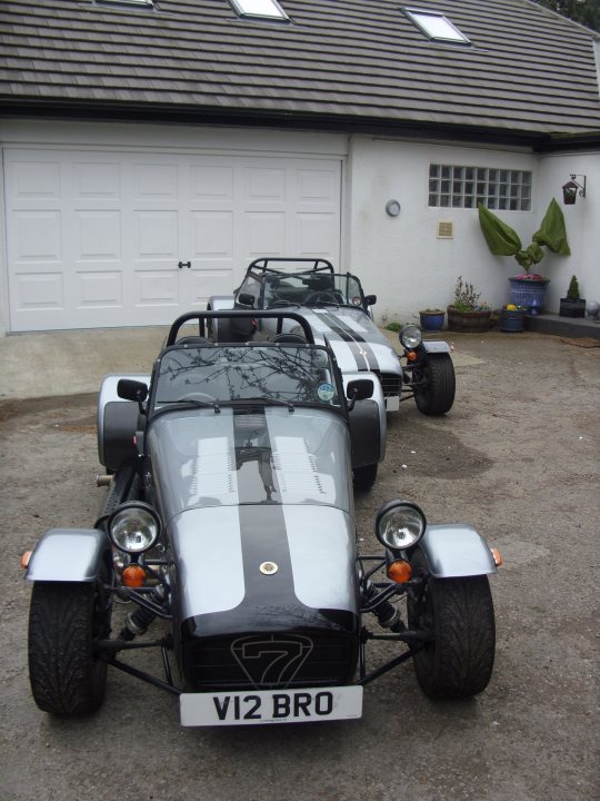 Livery Twin Pistonheads - The image features a garage with two classic sports cars parked outside, against a white house backdrop. Both cars are white with black stripes and have a vintage racing design. The cars are parked close to each other, and the license plate on the closer car reads "VI2 BRO". The garage door appears to be sturdy and insulated, typical of a garage designed to store valuable vehicles. The driveway is clean and well-maintained, with no visible trash or debris. The relative position of the cars suggests they are being displayed prominently.