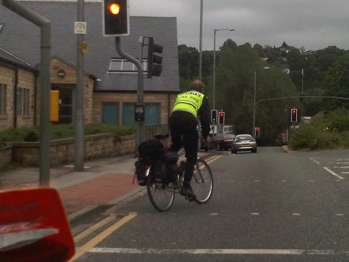 A man riding a bike down a street - Pistonheads - The image depicts a quiet residential street with traffic controls in place. A cyclist wearing a reflective jacket is seen mid-action, crossing the street and going against the traffic light. The cyclist is astride a bicycle and appears to be carrying a backpack. Other traffic participants include pedestrians and cars, and the sky is overcast with clouds, indicating a potentially gloomy or cool day.