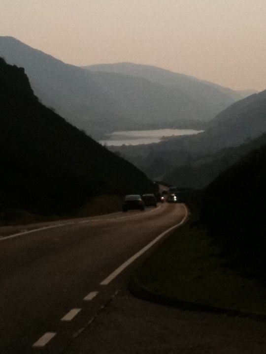 Pistonheads Blat Wales - The image showcases a serene mountain landscape, dominated by a winding road that curves along the base of the mountains. A few cars are driving on the road, their headlights illuminating the road ahead amidst the softly diffused evening light. The colors in the photo are dim, providing a calm and tranquil ambiance, with the setting sun casting a warm glow over the scene. The mountains in the background appear to be in a far distance, and their dark silhouettes contrast with the lighter tones of the sky and the road. The overall scene suggests a peaceful evening drive in a mountainous region.